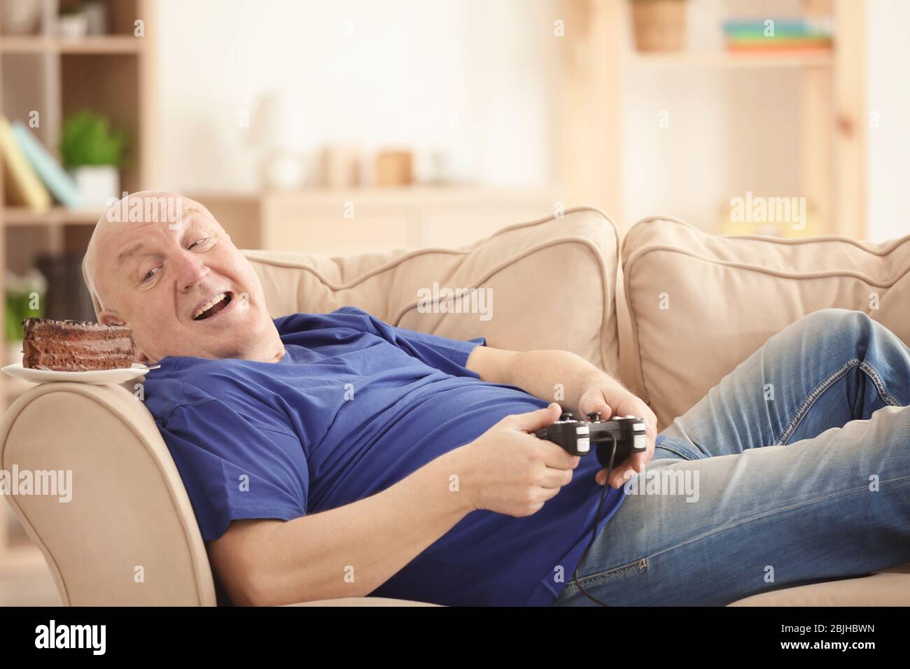 Uomo senior fat con console videogiochi e torta sdraiata sul divano a casa. Stile di vita sedentario Foto Stock