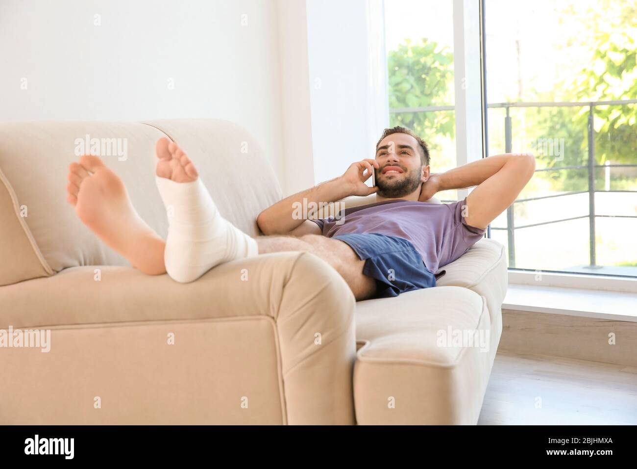 Bel giovane uomo con gamba a fascia che parla con il telefono cellulare mentre riposano sul divano a casa Foto Stock