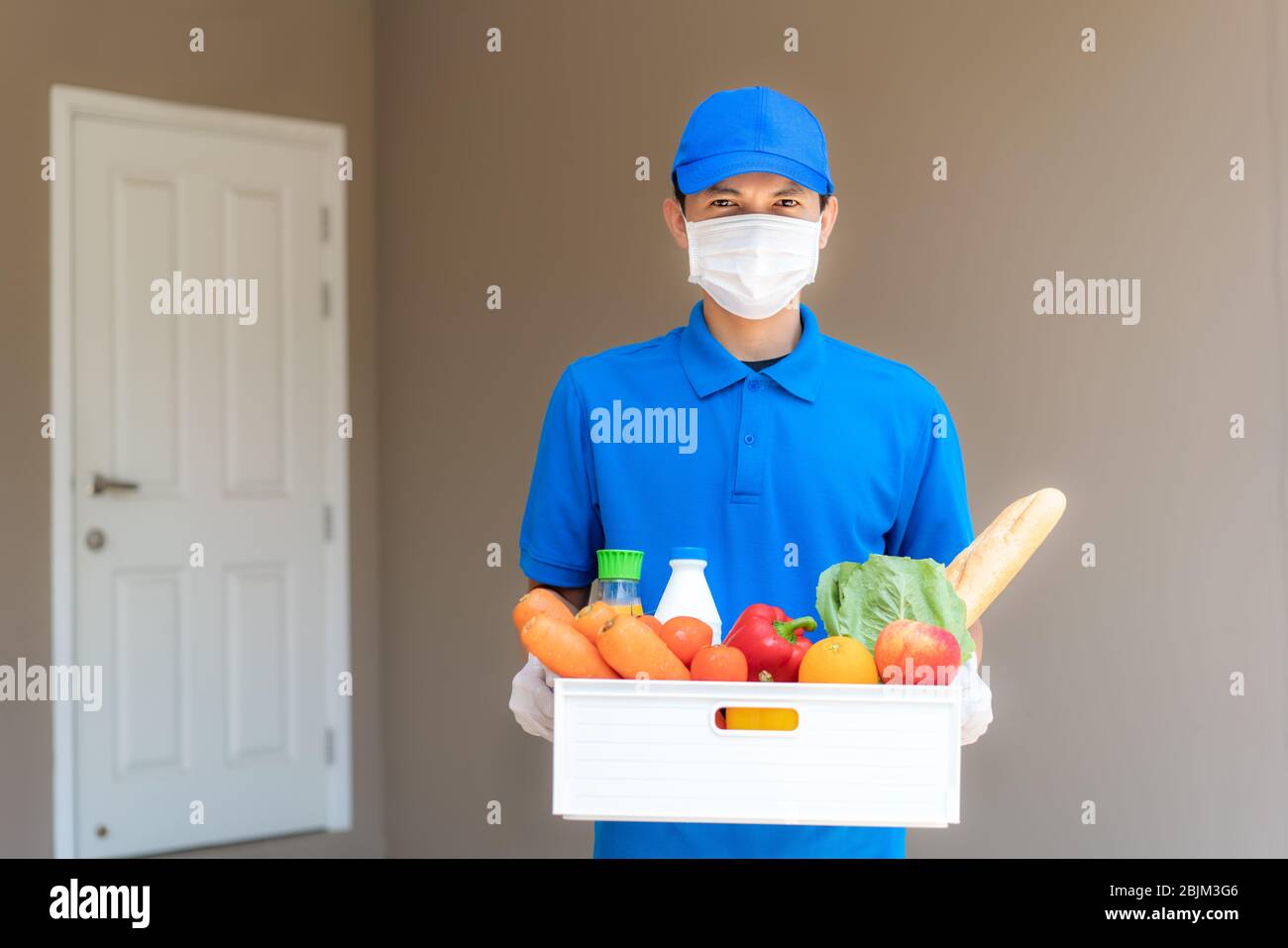 Uomo asiatico che indossa maschera facciale e guanto con scatola di generi alimentari di cibo, frutta, verdura e bevande davanti alla casa del cliente durante la t Foto Stock