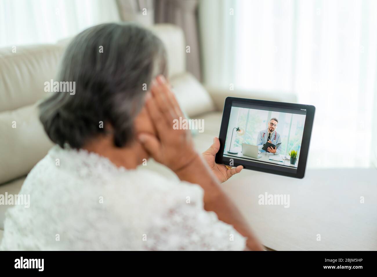 Vista posteriore della donna anziana che fa videochiamata con il suo medico con la sua sensazione di mal di testa sul tablet digitale online assistenza sanitaria tecnologia digitale Foto Stock
