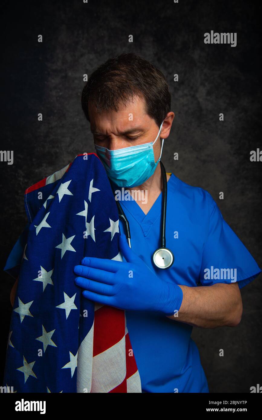 Medico maschile triste/pensivo in scrub blu ospedale con maschera facciale e stetoscopio, nutrendo la bandiera americana Stars & Stripes vicino al suo petto. Foto Stock