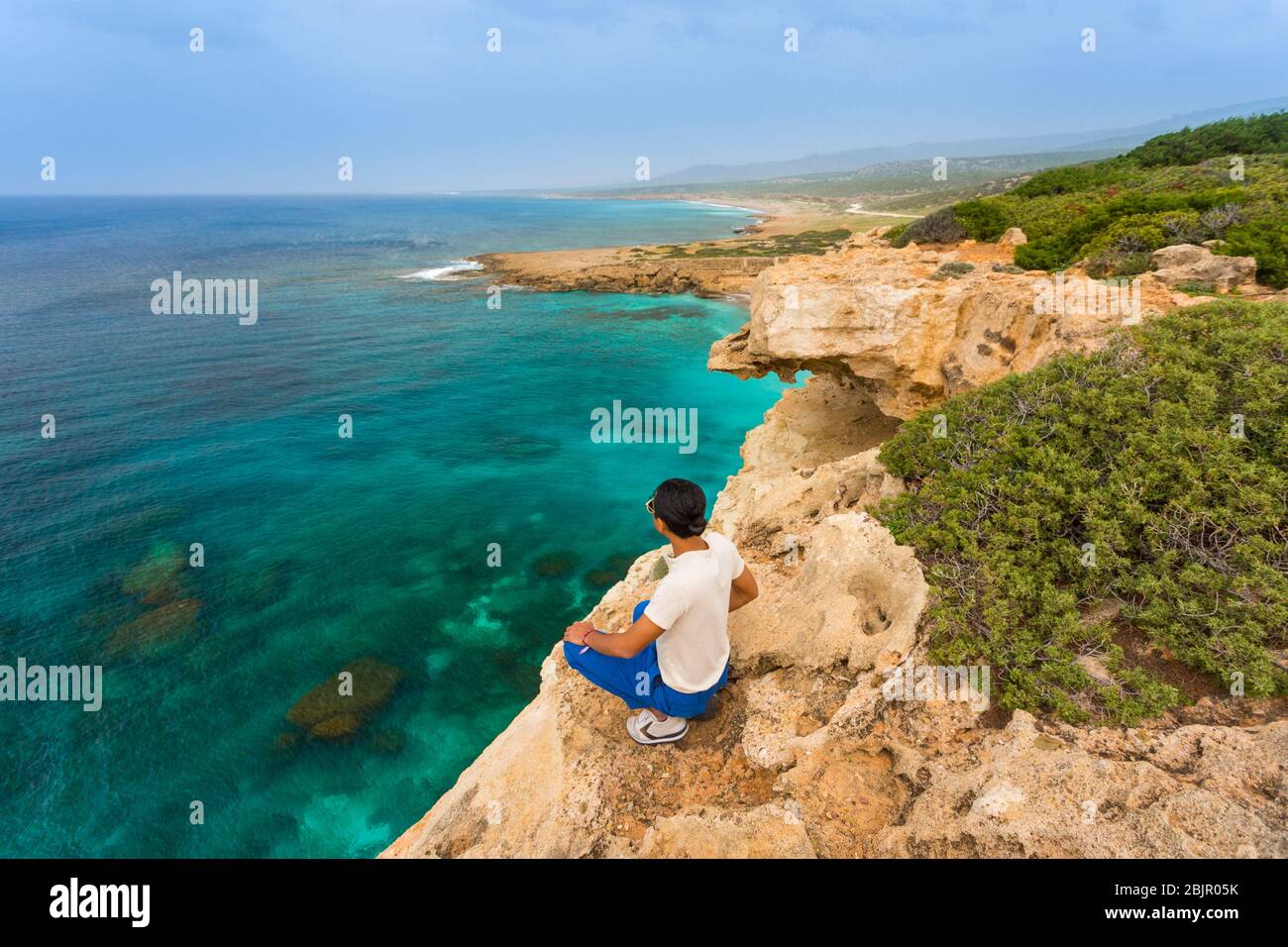 Giovane seduto sul bordo di una scogliera che si affaccia sull'oceano a Lara Beach sulla penisola di Akamas, Cipro Foto Stock