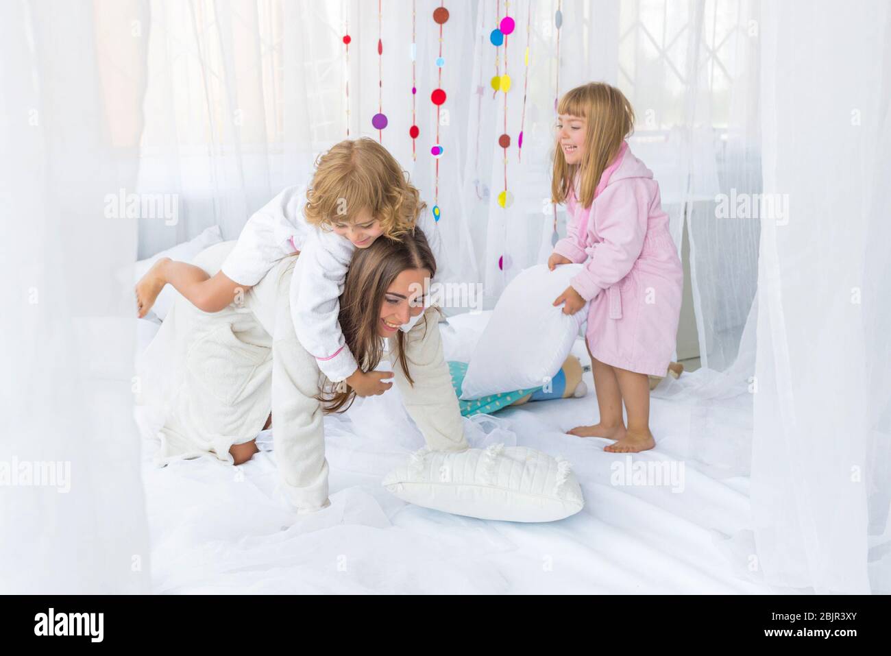 La giovane donna si diverte con i bambini che giocano con i cuscini sul letto della camera da letto. Soggiorno a casa e concetto di famiglia. Foto Stock