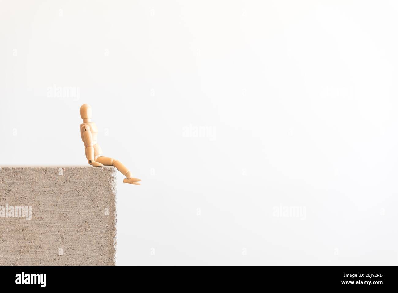 Figura umana in legno seduta sul bordo di un blocco di cemento, da sola. Simbolo di distanza sociale, ma anche di guardare avanti Foto Stock