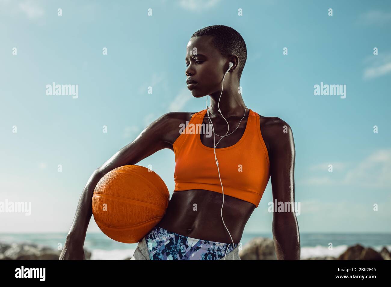 Donna africana in abbigliamento sportivo che tiene un basket in piedi alla spiaggia. Donna fitness che indossa gli auricolari per ascoltare musica dopo l'allenamento sportivo. Foto Stock