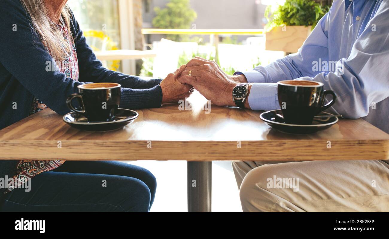 Un colpo corto di una coppia anziana seduta ad un tavolo della caffetteria tenendo le mani con due tazze di caffè davanti. Primo piano di uomo e donna con le mani in mano Foto Stock