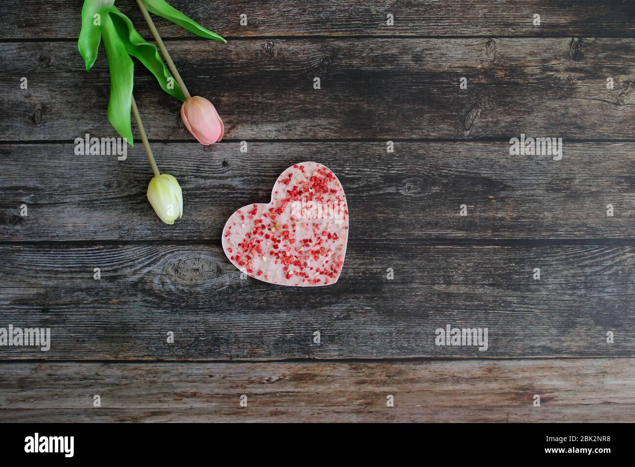 Un cioccolato rosa a forma di cuore con fragole essiccate su un tavolo vintage di legno scuro con fiori di tulipano. Concetto di amore e biglietto di auguri. Foto Stock