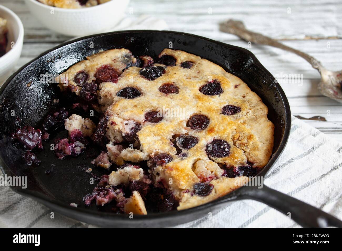 Metà mangiata fresca fatta in casa ciottoli di ciliegia cotti in una padella di ghisa su un rustico tavolo di legno bianco. Messa a fuoco selettiva con sfondo sfocato. Foto Stock