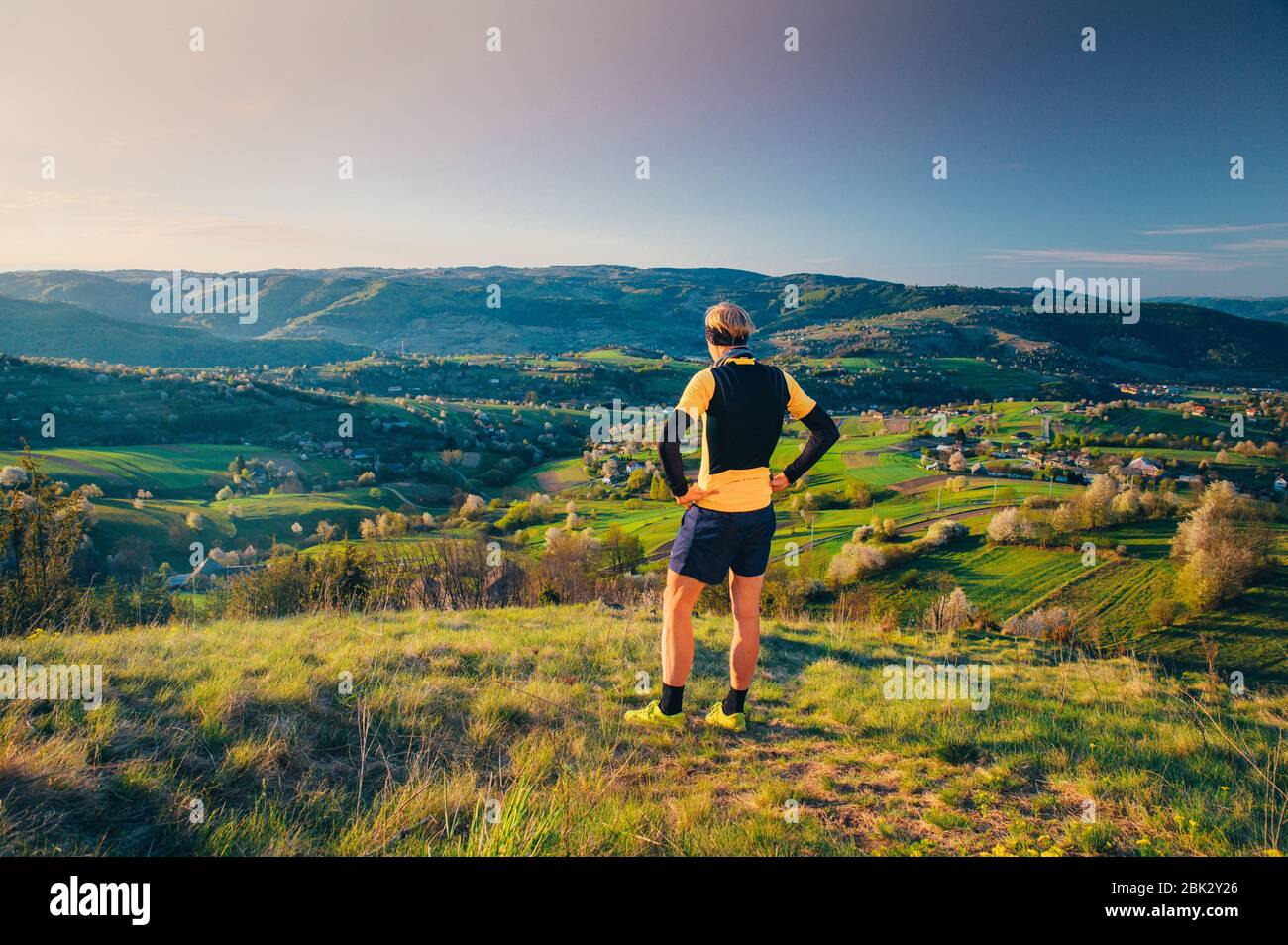 Atleta maschile in piedi sulla cima della collina e guardando il bellissimo paesaggio rurale primavera in luce alba. Esercizio mattutino, foto dello sport di corsa. Foto Stock