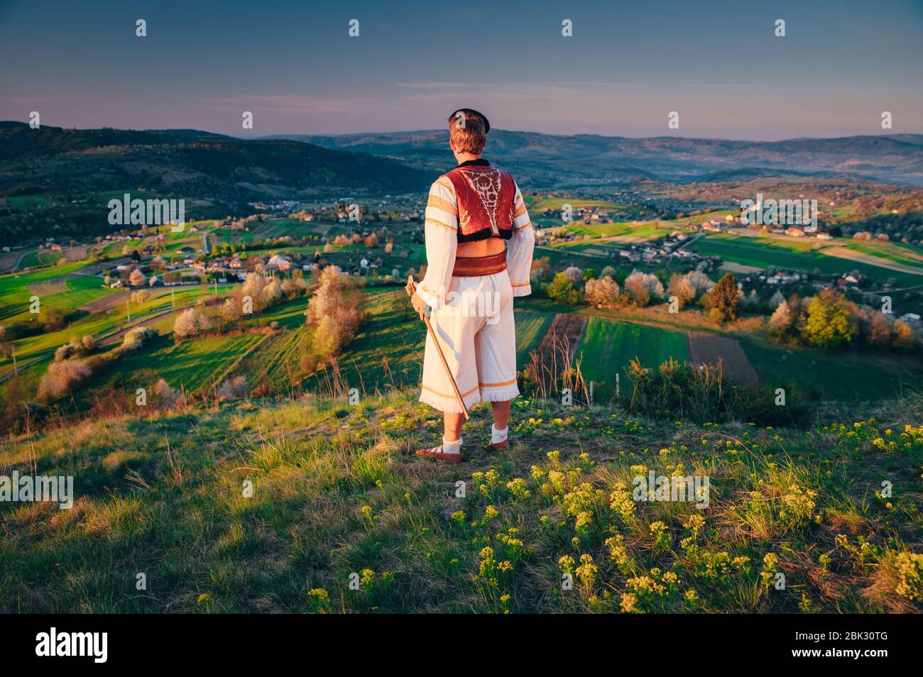 Un giovane in costume popolare slovacco guarda il paesaggio primaverile nel villaggio di Hrinova in Slovacchia. Sole che sorge e alberi in fiore primavera nel Foto Stock