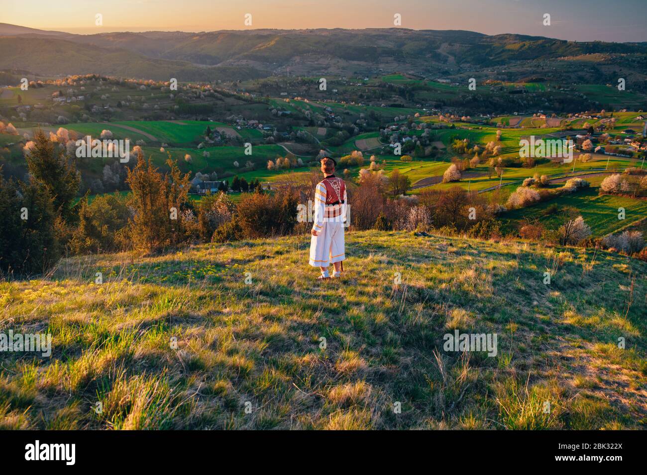 Un giovane in costume popolare slovacco guarda il paesaggio primaverile nel villaggio di Hrinova in Slovacchia. Sole che sorge e alberi in fiore primavera nel Foto Stock
