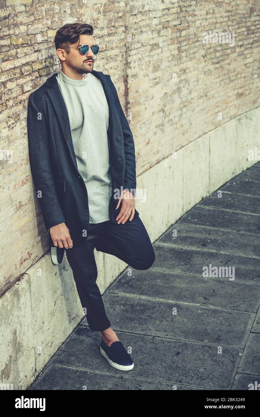 Bell'uomo elegante appoggiato su una parete con occhiali da sole e  acconciatura moderna e barba. Esterno in strada. T-shirt bianca e giacca  nera Foto stock - Alamy