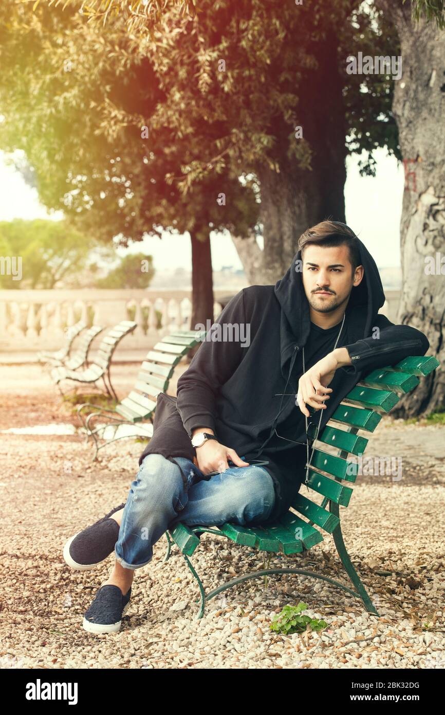 Giovane e alla moda, rilassante su una panchina in un parco con alberi. Il ragazzo si veste di moda, indossando parzialmente una felpa con cappuccio. All'aperto in un parco. Foto Stock