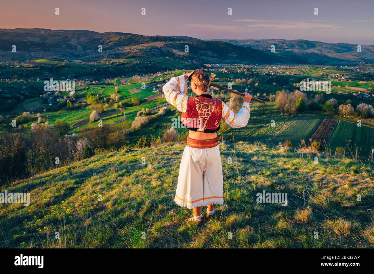 Uomo in tradizionale abito Folk guardando luce alba in bello caldo paesaggio primaverile. Prato rurale e fiori primaverili alberi di ciliegio sullo sfondo Foto Stock