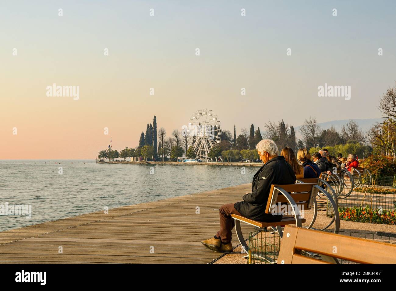 Anziani e altre persone seduti su panchine sulla riva del lago con una ruota panoramica sullo sfondo al tramonto, Bardolino, VeronaVeneto, Italia Foto Stock