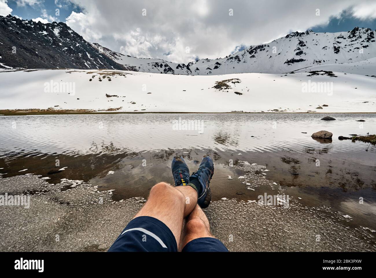 Gambe di uomo in scarpe di tracking e vista del ghiacciaio nevoso sul lago di montagna Foto Stock