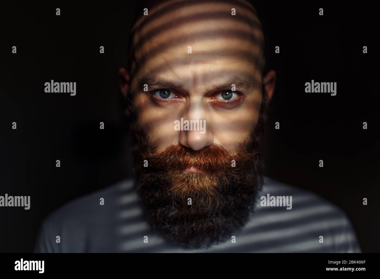 Ritratto ravvicinato di un uomo barbuto di mezza età con occhi espressivi e ombre a righe sul suo volto con sfondo nero Foto Stock