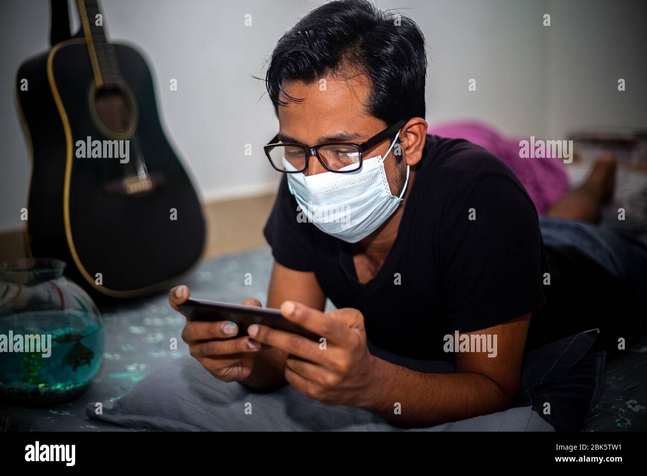 Un giovane uomo con maschera chirurgica stava giocando a giochi mobili a casa sua a causa della quarantena domestica del coronavirus a Dhaka. Foto Stock