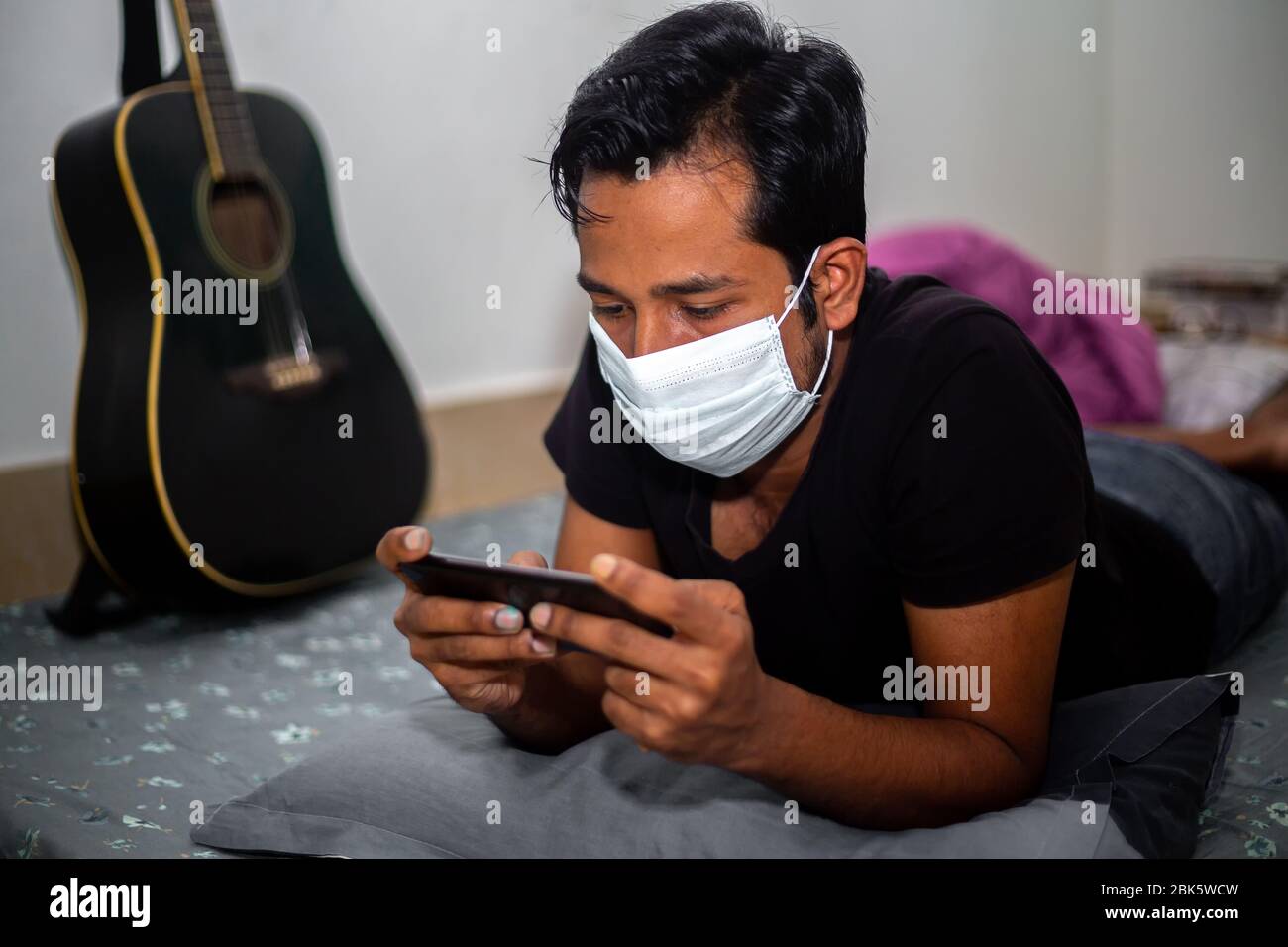 Un giovane uomo con maschera chirurgica stava giocando a giochi mobili a casa sua a causa della quarantena domestica del coronavirus a Dhaka. Foto Stock