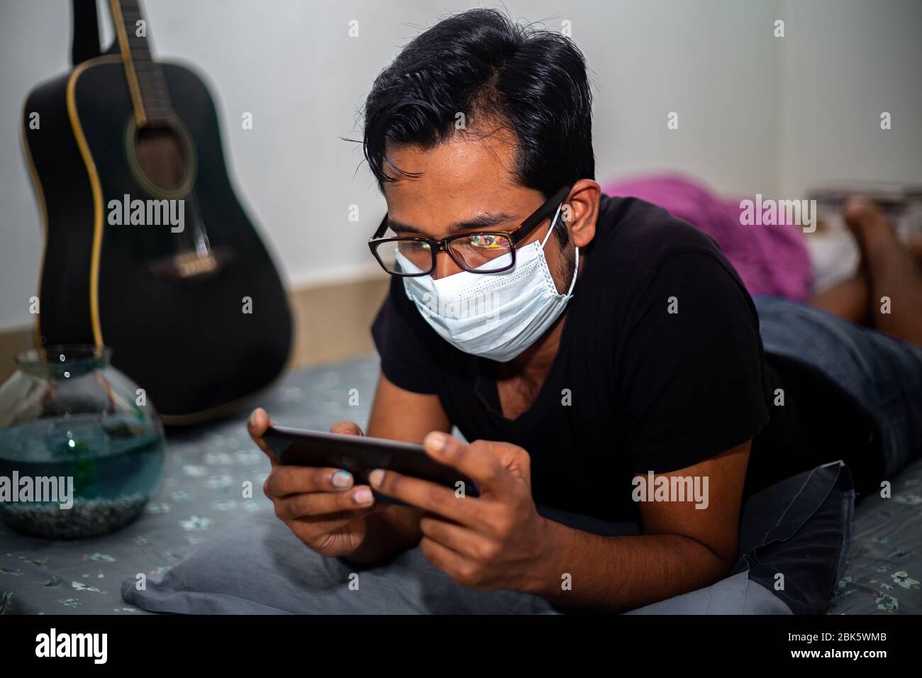 Un giovane uomo con maschera chirurgica stava giocando a giochi mobili a casa sua a causa della quarantena domestica del coronavirus a Dhaka. Foto Stock