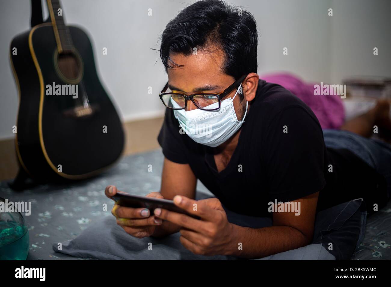 Un giovane uomo con maschera chirurgica stava giocando a giochi mobili a casa sua a causa della quarantena domestica del coronavirus a Dhaka. Foto Stock