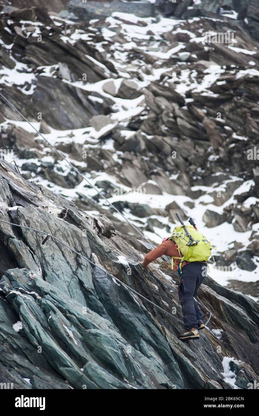 Vista posteriore dell'alpinista con zaino in corda mentre si sale sul crinale alpino. Uomo arrampicatore ascendente scogliera di montagna, cercando di raggiungere la cima della montagna. Concetto di alpinismo, alpinismo e arrampicata alpina. Foto Stock