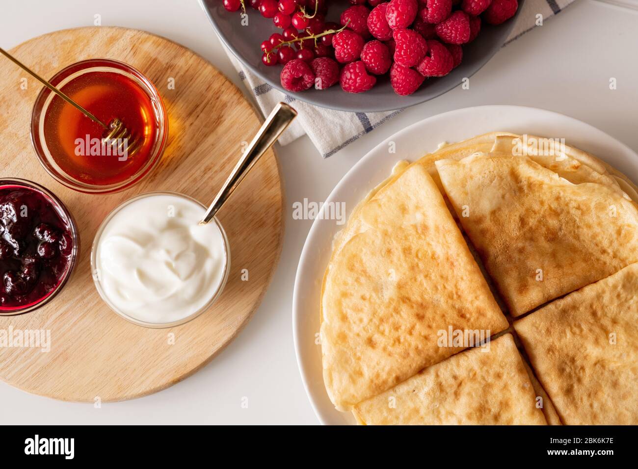 Disposizione piatta di quattro pancake piegati sul piatto, lamponi freschi e ribes rosso, piccole ciotole di vetro con miele, marmellata di ciliegie e crema di caffè a bordo Foto Stock