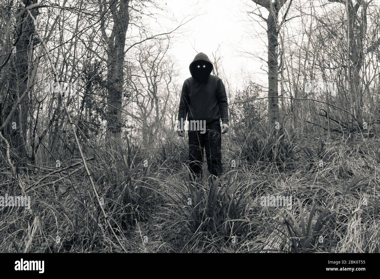 Una figura spaventosa e con cappuccio, con occhi malvagi e spazi neri vuoti dove dovrebbe essere il suo volto, in piedi in una foresta in inverno. Con una modifica vintage. Foto Stock