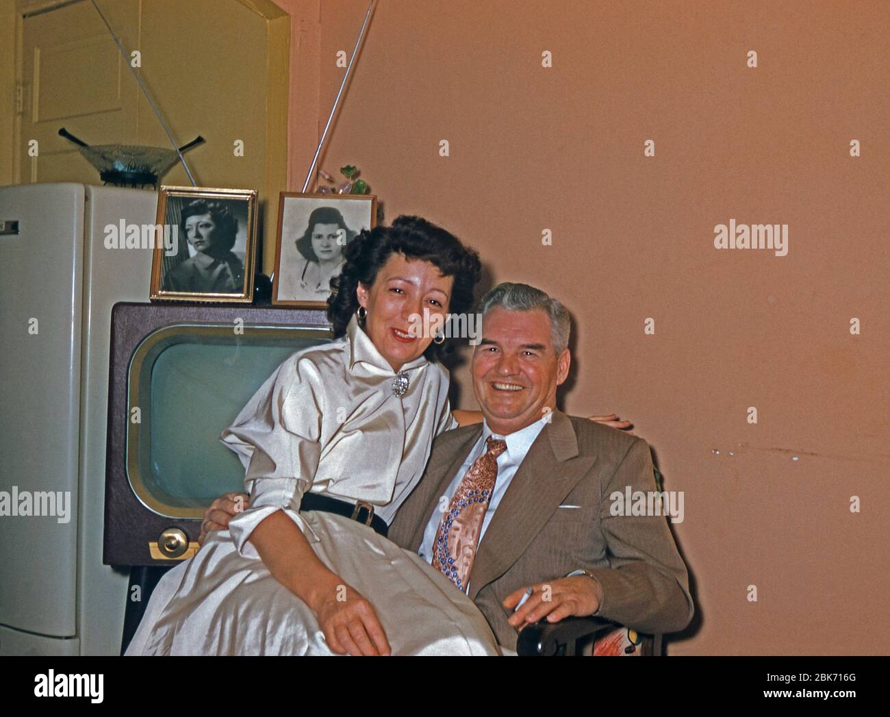 Una coppia felice più anziana nella loro casa, USA, c 1955. La donna si siede sul ginocchio dell'uomo mentre tiene una sigaretta. Un televisore e un frigorifero sono dietro di loro in una stanza dipinta nei colori tipici di quell'epoca. Questa immagine proviene da un vecchio lucido a colori americano dilettante Kodak. Foto Stock