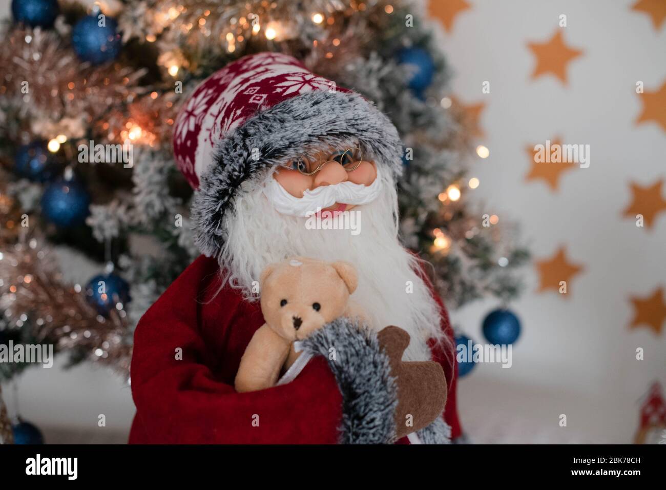 Bambola Babbo Natale, grande personaggio di stand natalizio che tiene un simpatico orsacchiotto, posizionato di fronte a un albero di Natale illuminato, decorazione festosa per le vacanze Foto Stock