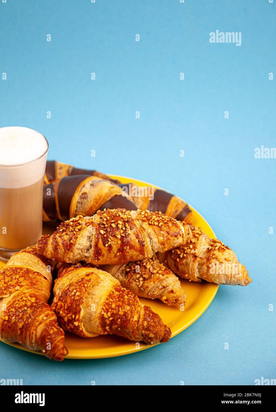 Croissant su una pelle di vitello gialla. Sfondo blu con posto per il testo. Un bicchiere alto con una bevanda calda del mattino - caffè latte con latte. Appetitoso Foto Stock