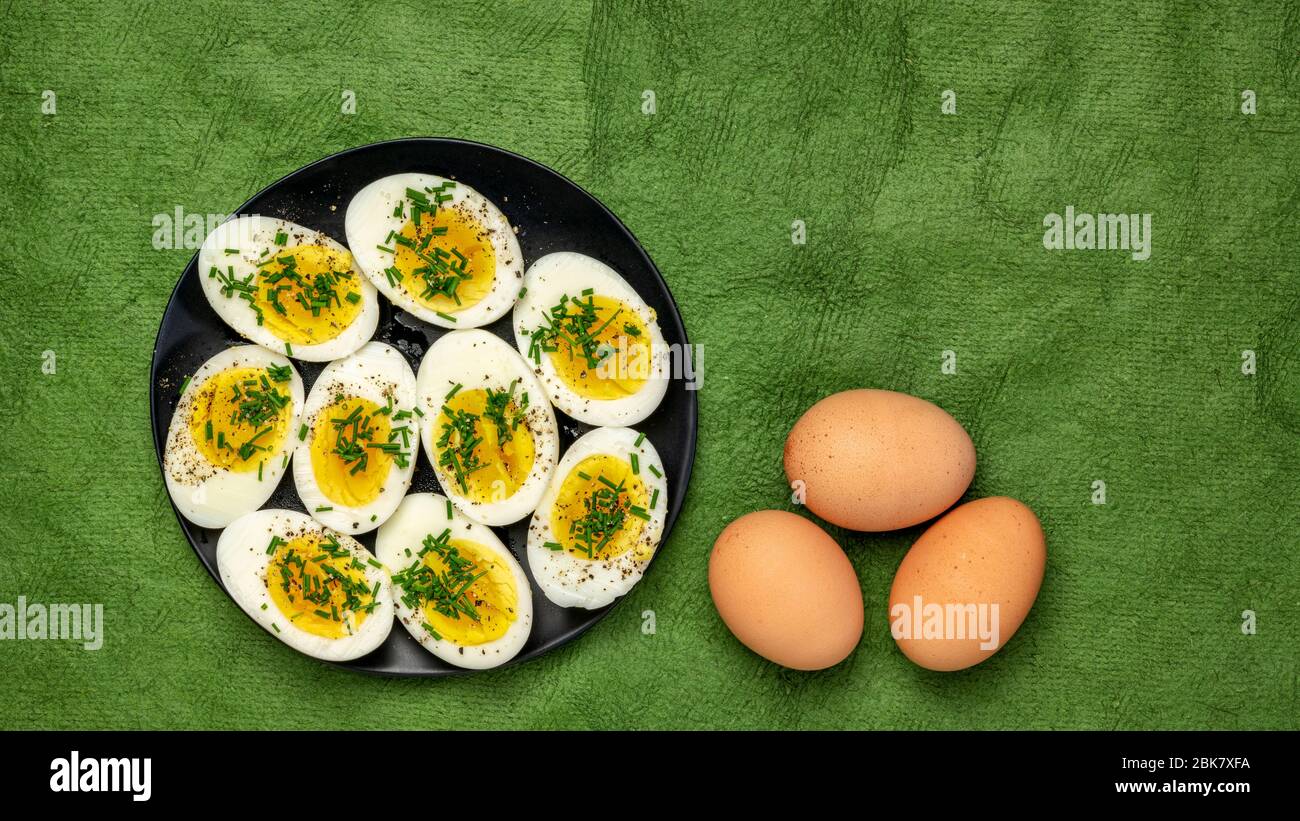Uova di pollo bollite con erba cipollina fresca verde, sale e pepe su un piatto nero contro carta testurizzata fatta a mano - Pasqua o primavera concetto Foto Stock