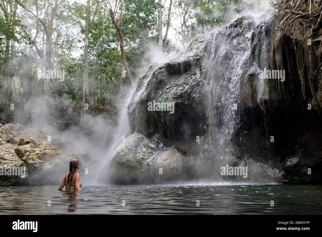 Donna in bikini che fa un bagno in un lago dove cade una cascata di sorgenti calde fumanti. Foto Stock