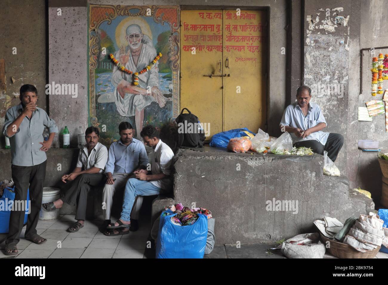 Uomini a Mumbai, Maharashtra, India, passando il tempo in un sottopassaggio scuro di fronte ad un quadro ghirlang del 19 ° secolo indù san Sai Baba di Shirdi Foto Stock