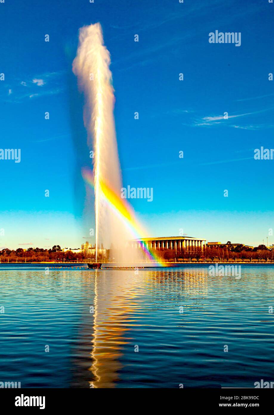 Rainbow through Captain Cook Memorial Water Jet sul lago Burley Griffin a Canberra, la capitale nazionale australiana con la National Library in background Foto Stock