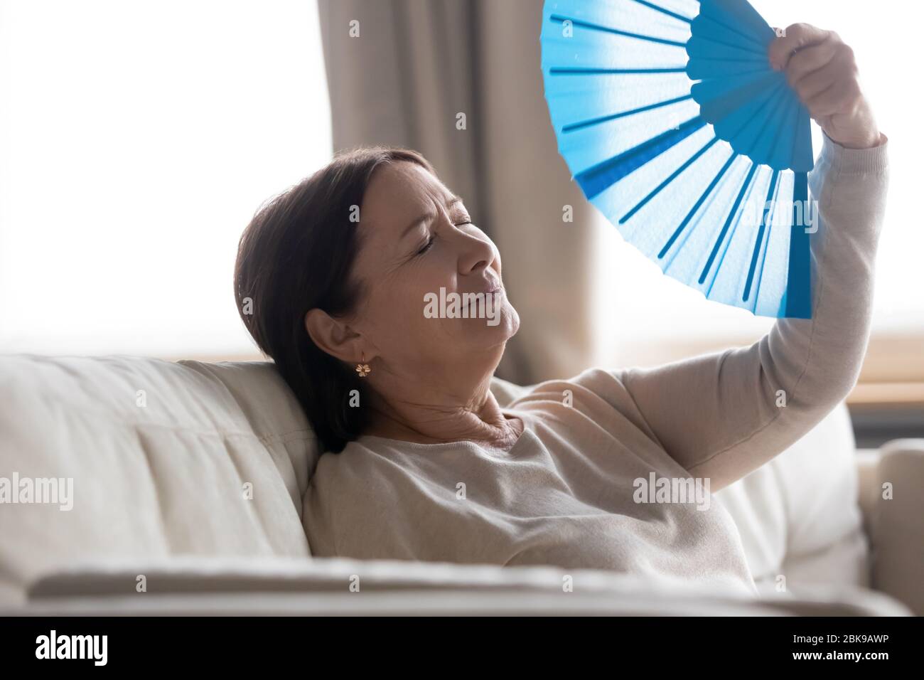 Donna stanca di mezza età che agita il ventilatore blu, soffrendo dal calore Foto Stock