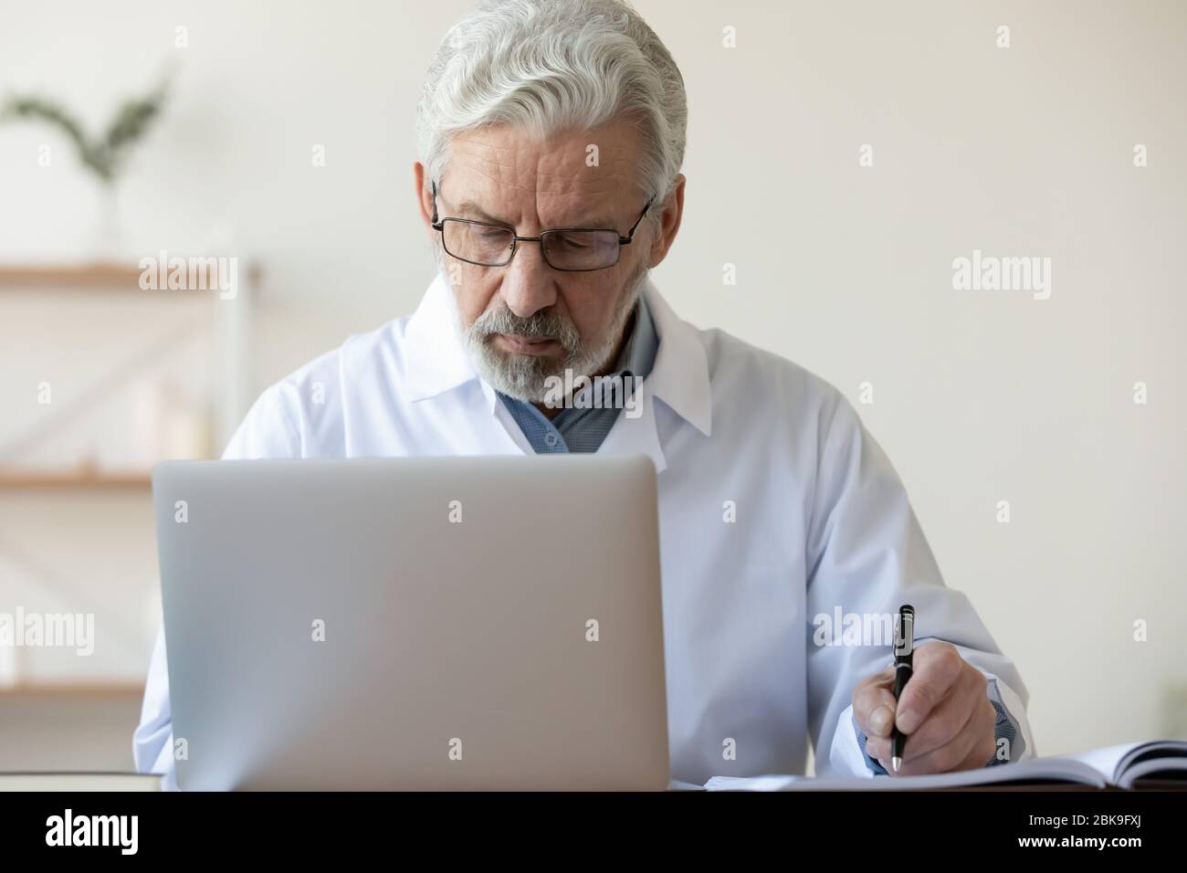 Un vecchio medico serio che scriveva appunti, usando il laptop in un ufficio medico Foto Stock
