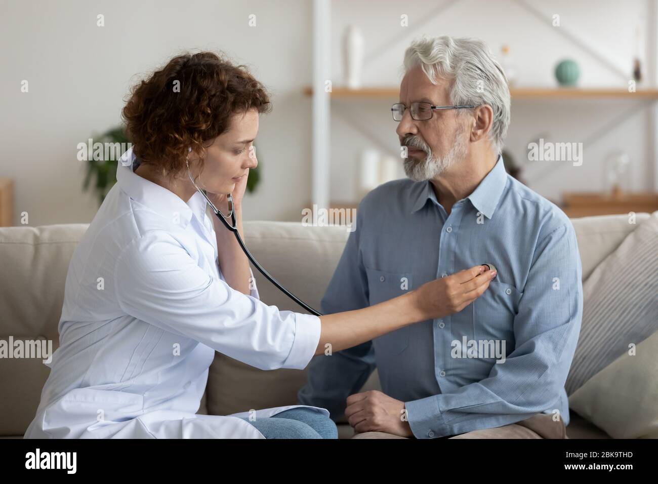 Medico femminile che tiene stetoscopio in ascolto paziente anziano durante la visita di homecare Foto Stock