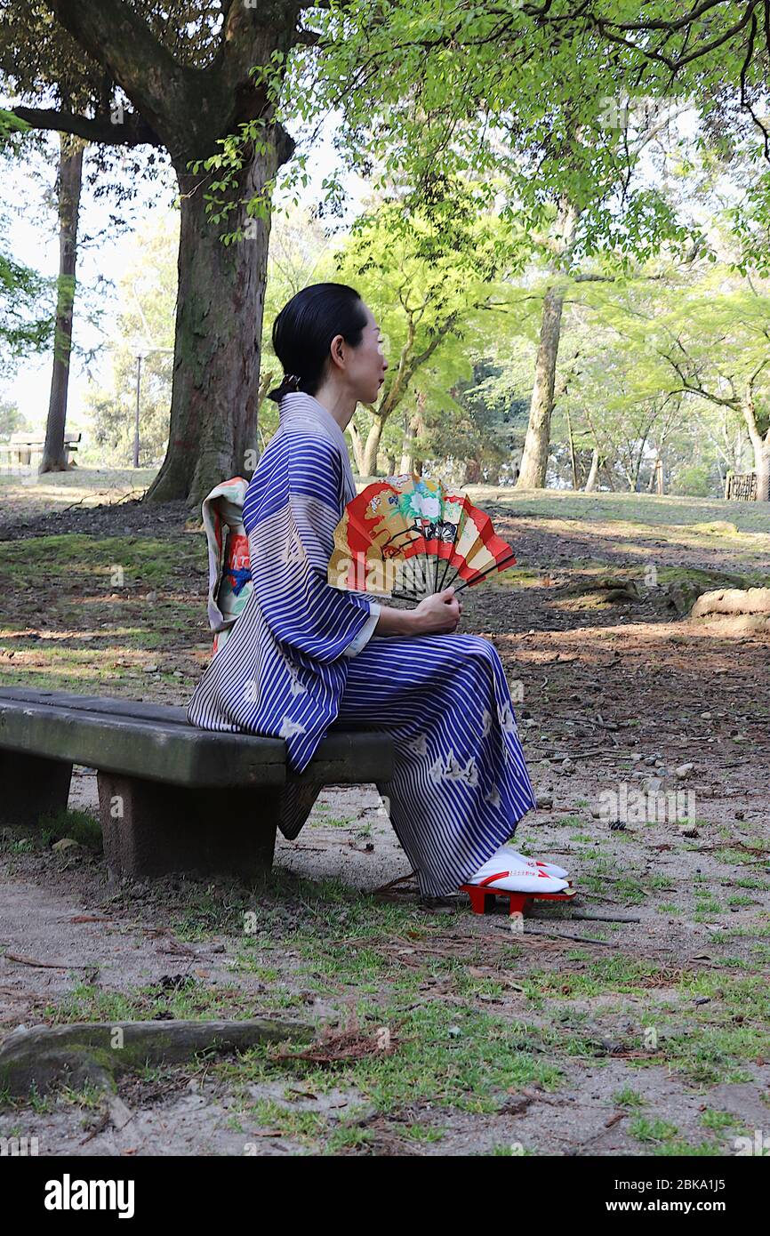 Donna giapponese in kimono con un ventilatore in mano seduto su una panchina in un parco Foto Stock