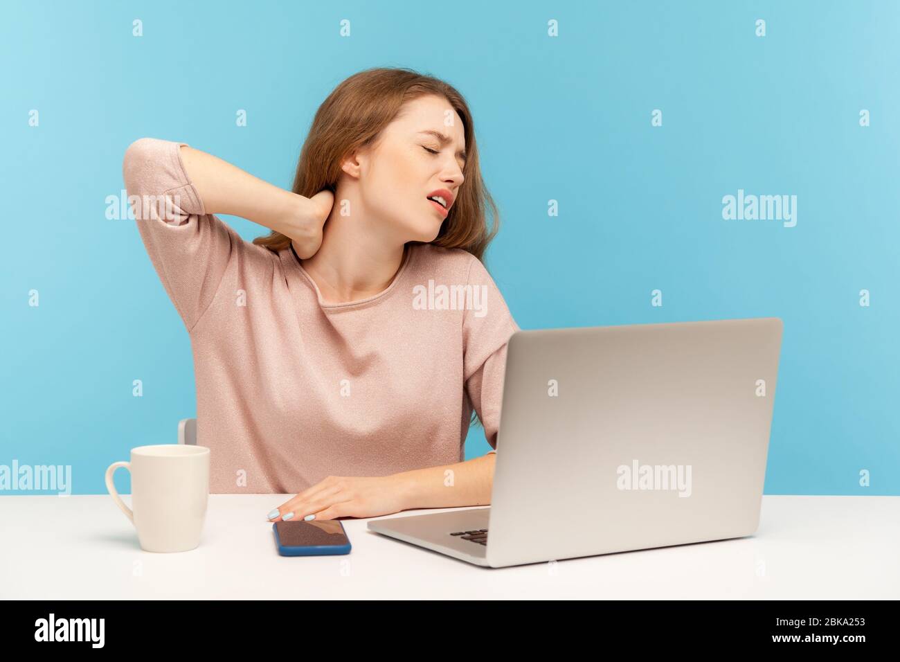 Giovane donna malata che lavora sul posto di lavoro con il laptop che si sente malato con il collo irritato, ferisce spalle e schiena, massaggiando dolorosi muscoli rigidi. Foto Stock