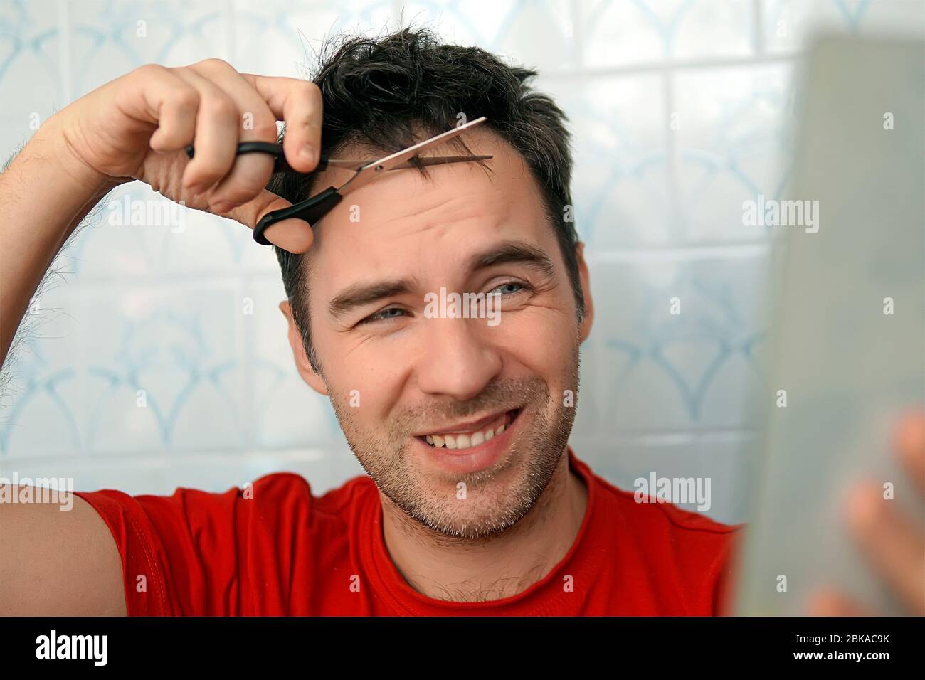 Bell'uomo che taglia i propri capelli con una forbici e guarda nello specchio. Autosocura nelle condizioni di quarantena globale e acconciatori chiusi An Foto Stock