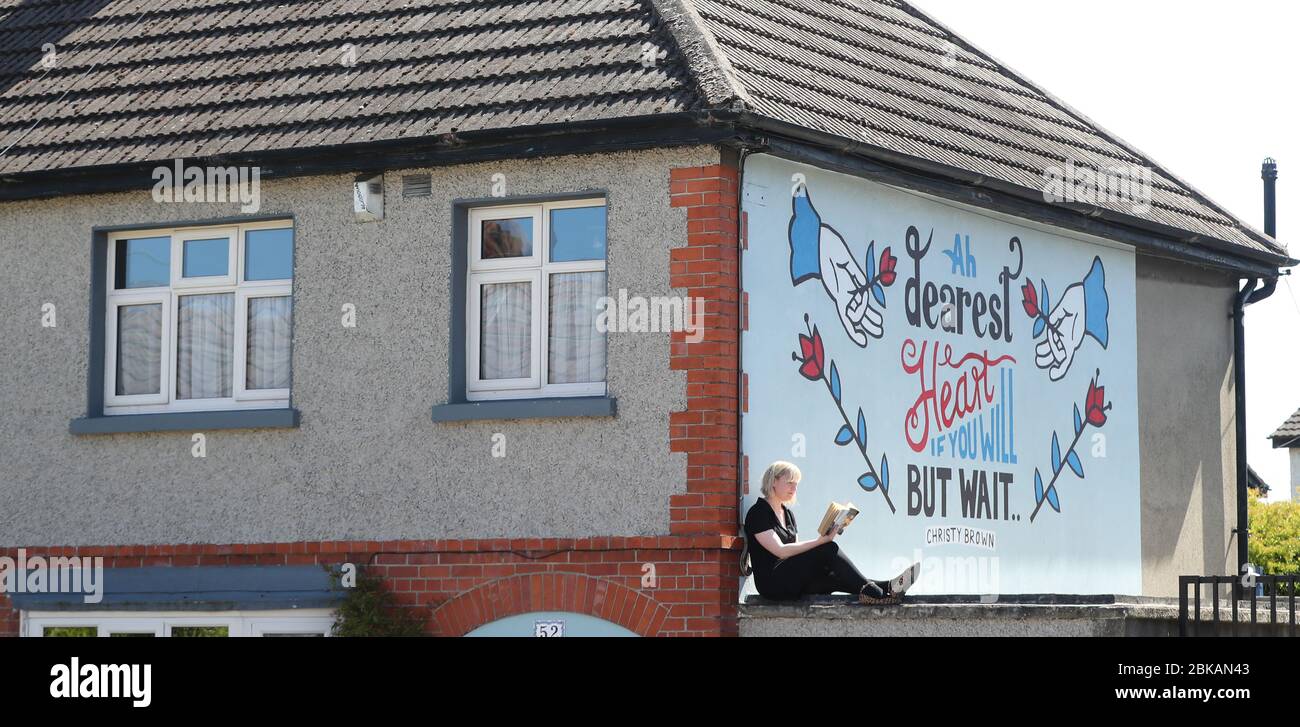 L'artista Sarah Bracken Soper legge una copia del mio piede sinistro da Christy Brown sotto un murale che ha dipinto citando l'autore sul lato della sua casa a Crumlin Dublino durante il Covid-19 Lockdown. Foto Stock