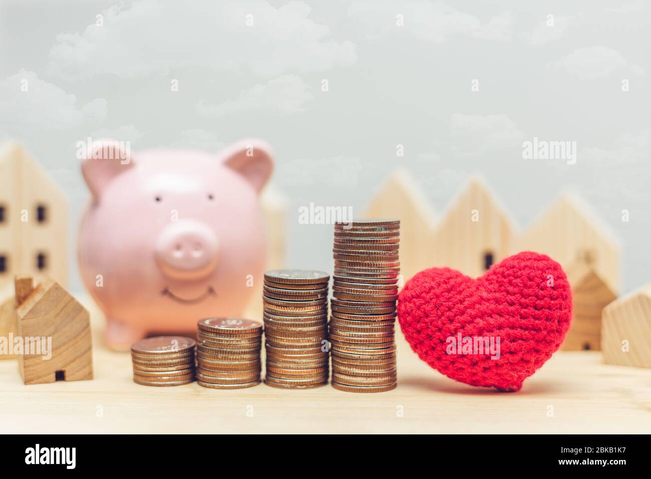Pila di monete con modello domestico di legno e banca del piggy per i soldi di risparmio per comprare una nuova sede con il concetto di cuore di amore. Foto Stock