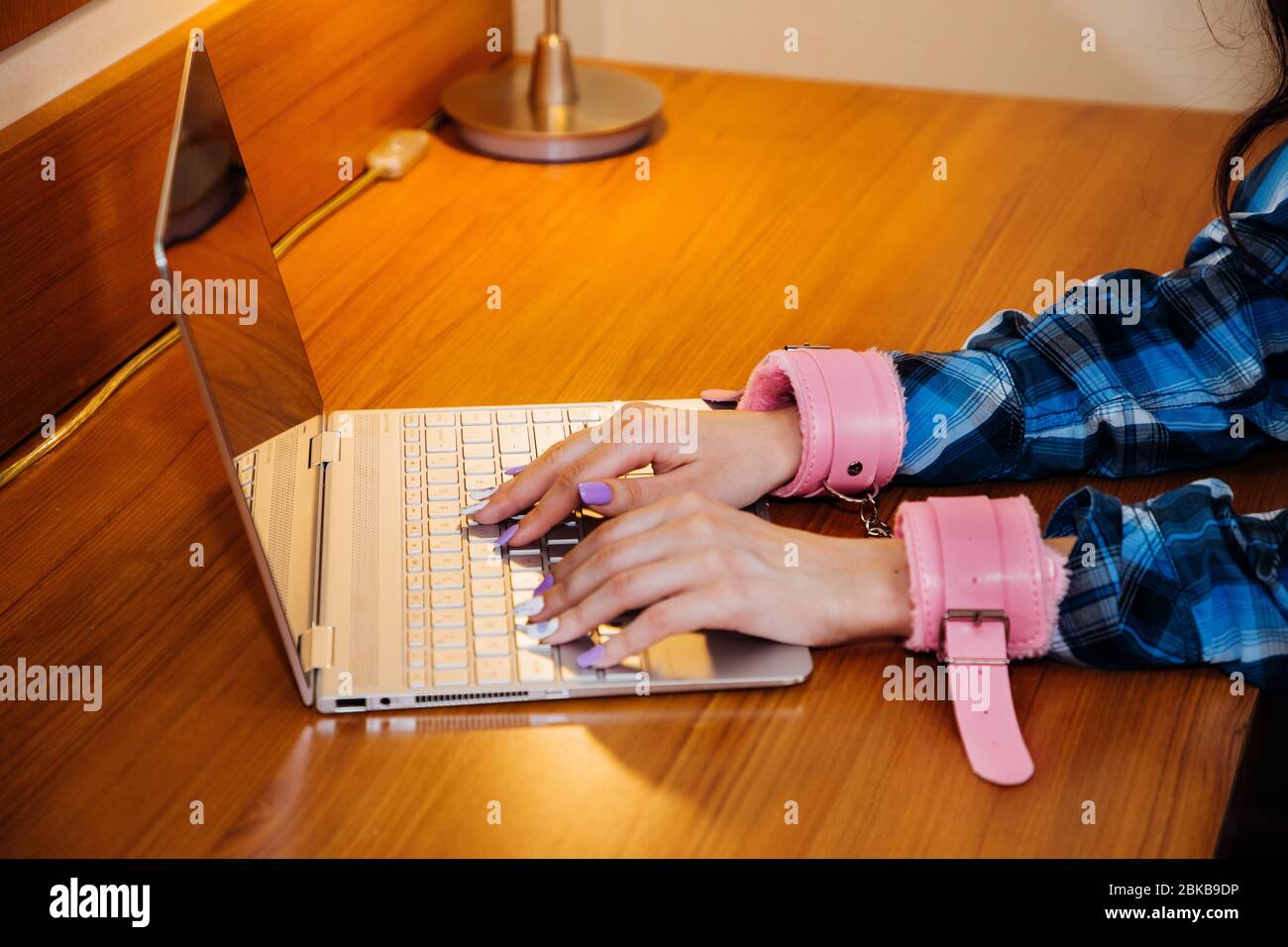 Le mani con manette rosa scrivono sulla tastiera di un notebook sottile; simbolo per essere attaccate al computer durante la quarantena, lo stress o la dipendenza Foto Stock