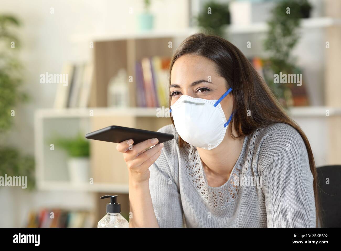 Donna felice che utilizza il sistema di riconoscimento vocale sullo smartphone in confino-19 maschera che indossa seduto a casa Foto Stock