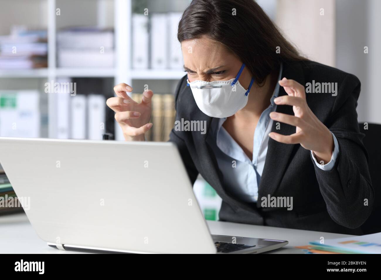 Donna esecutiva arrabbiata che legge le cattive notizie sul laptop evitando covid-19 con maschera su una scrivania in ufficio Foto Stock