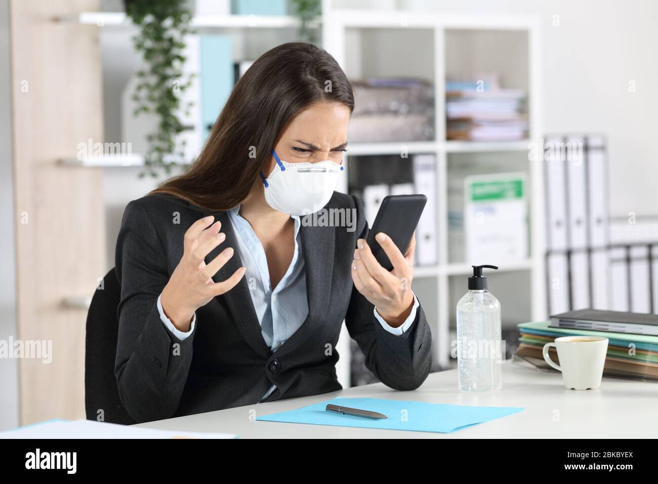 Donna executive arrabbiata che cerca smartphone evitando covid-19 con maschera seduta su una scrivania in ufficio Foto Stock