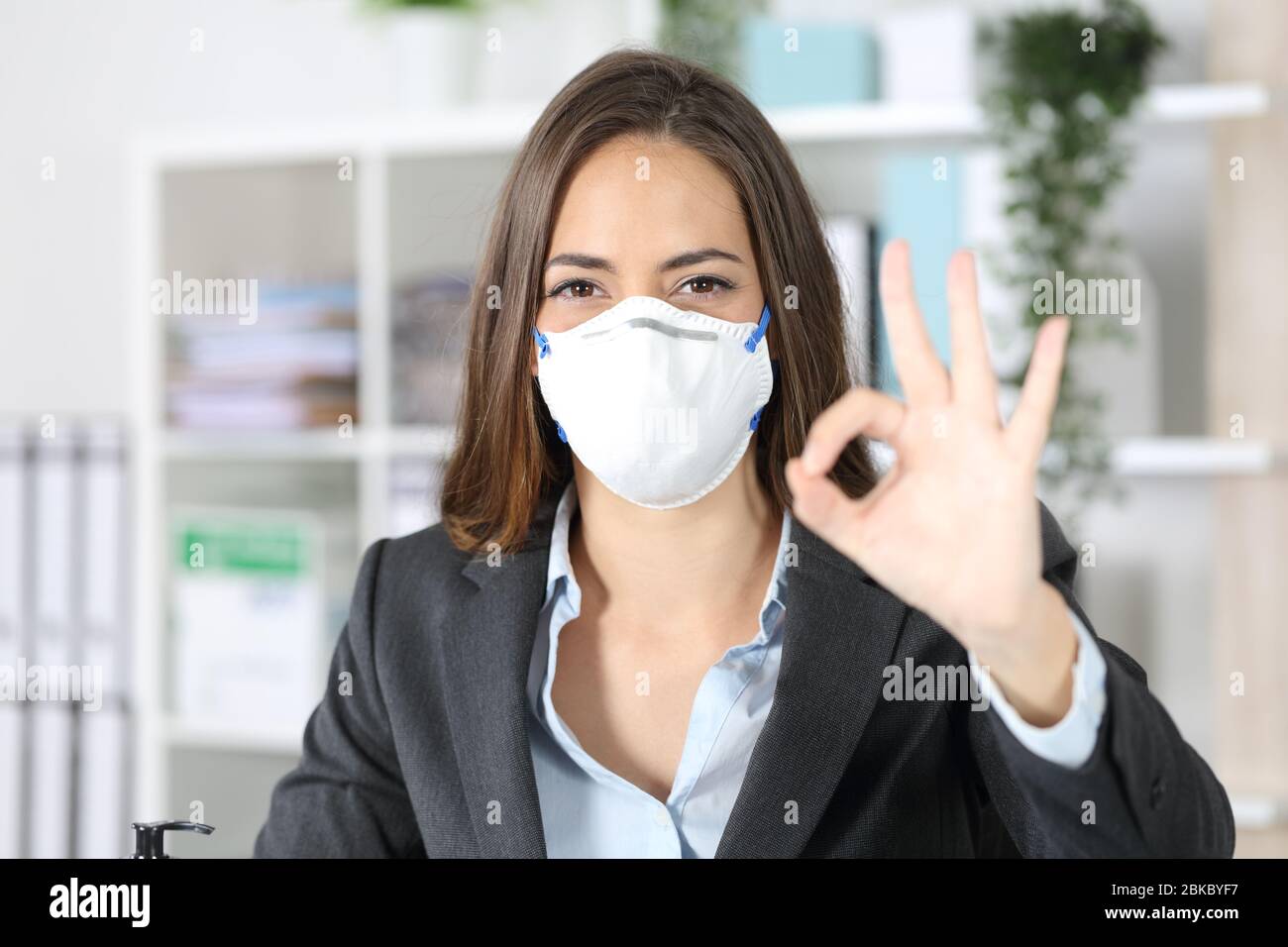 Vista frontale della donna executive gesturing ok evitando coronavirus guardare fotocamera con maschera protettiva in ufficio Foto Stock