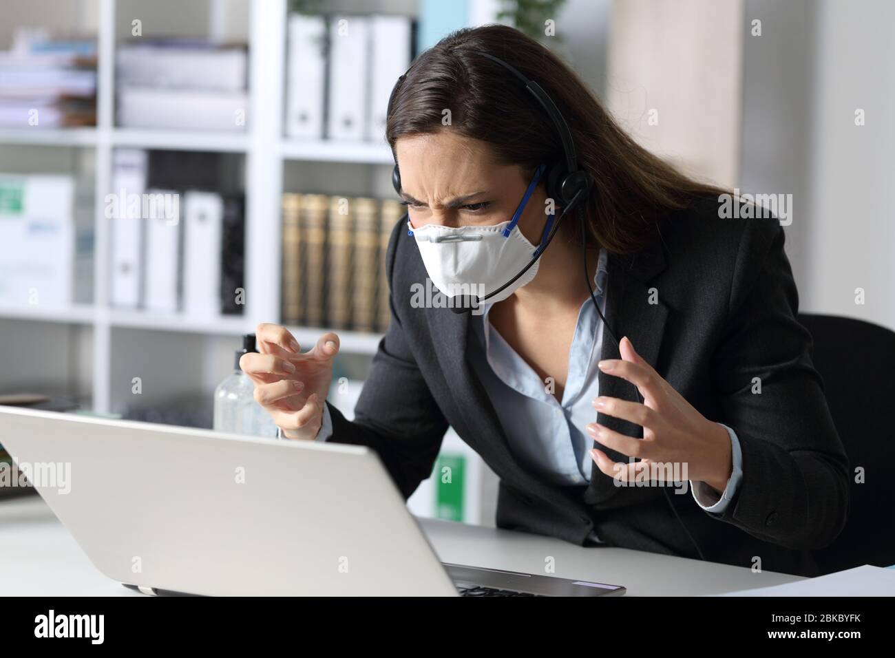Donna di telemarketer arrabbiata che cerca un notebook evitando covid-19 con maschera seduta su una scrivania in ufficio Foto Stock