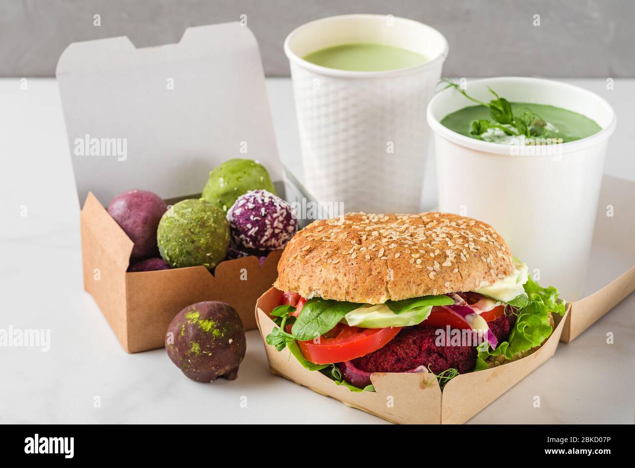 Consegna di cibo. Hamburger di barbabietola vegan e palle energetiche in scatole di carta ecologica con zuppa di panna di piselli e latte opaco in tazze monouso. Pranzo con dieta sana per la ta Foto Stock